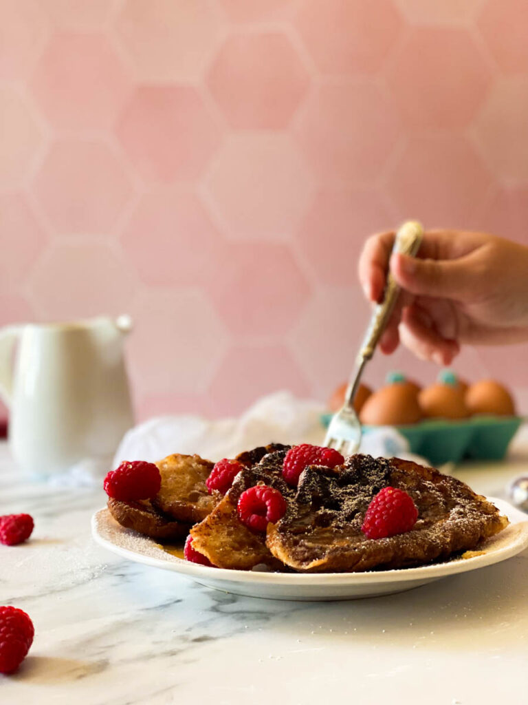 hand reaching with a fork for a croissant with raspberries, in the back, eggs, towel and milk jug