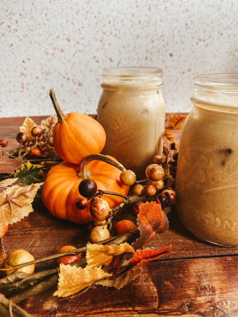 Pumpkin Spice Latte Iced Easy Recipe 2 This image shows two mason jars filled with pumpkin spice latte iced, set on a rustic wooden surface. Surrounding the jars are small decorative pumpkins, autumn leaves, and berries, creating a cozy fall atmosphere. The background features a speckled white surface, adding a warm and inviting seasonal touch to the scene.