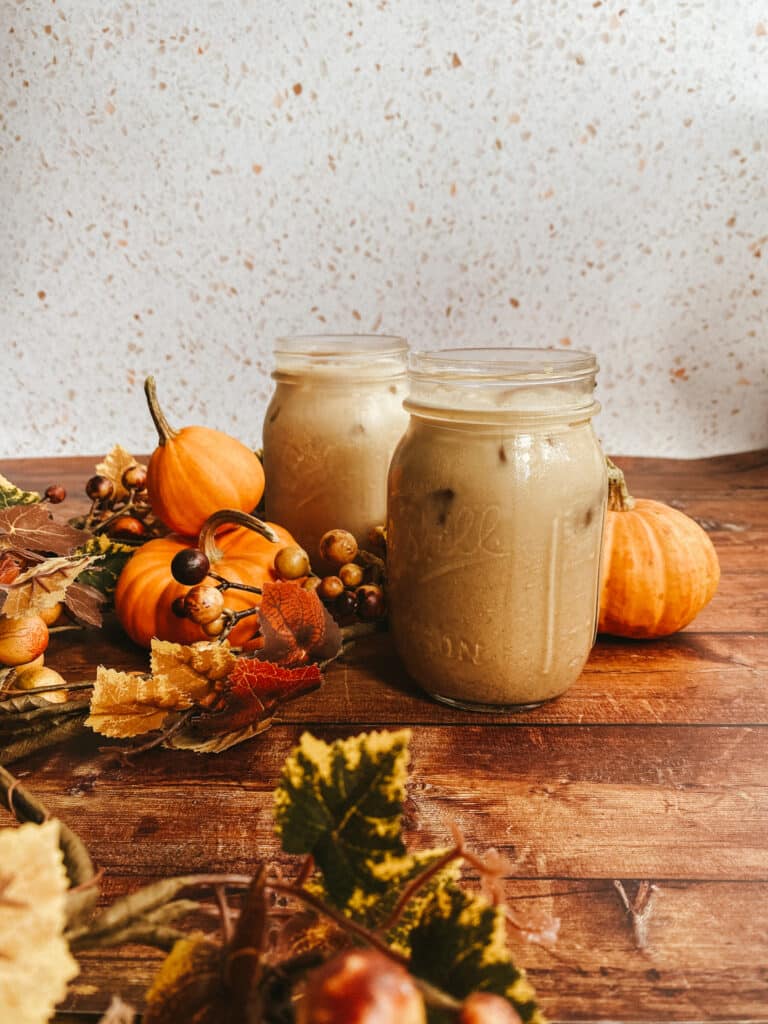 Pumpkin Spice Latte Iced Easy Recipe 9 This image shows two mason jars filled with pumpkin spice latte iced, set on a rustic wooden surface. Surrounding the jars are small decorative pumpkins, autumn leaves, and berries, creating a cozy fall atmosphere. The background features a speckled white surface, adding a warm and inviting seasonal touch to the scene.