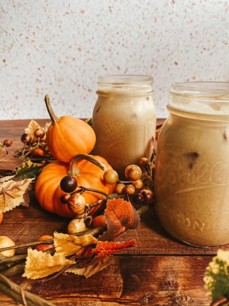 Pumpkin Spice Latte Iced Easy Recipe 1 This image shows two mason jars filled with pumpkin spice latte iced, set on a rustic wooden surface. Surrounding the jars are small decorative pumpkins, autumn leaves, and berries, creating a cozy fall atmosphere. The background features a speckled white surface, adding a warm and inviting seasonal touch to the scene.