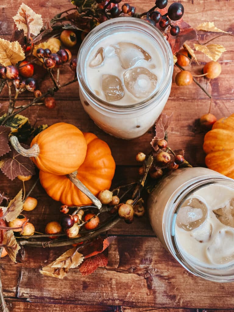 Pumpkin Spice Latte Iced Easy Recipe 8 This image shows two mason jars filled with pumpkin spice latte iced, set on a rustic wooden surface. Surrounding the jars are small decorative pumpkins, autumn leaves, and berries, creating a cozy fall atmosphere. The background features a speckled white surface, adding a warm and inviting seasonal touch to the scene.