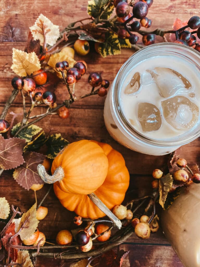 Pumpkin Spice Latte Iced Easy Recipe 3 This image shows two mason jars filled with pumpkin spice latte iced, set on a rustic wooden surface. Surrounding the jars are small decorative pumpkins, autumn leaves, and berries, creating a cozy fall atmosphere. The background features a speckled white surface, adding a warm and inviting seasonal touch to the scene.