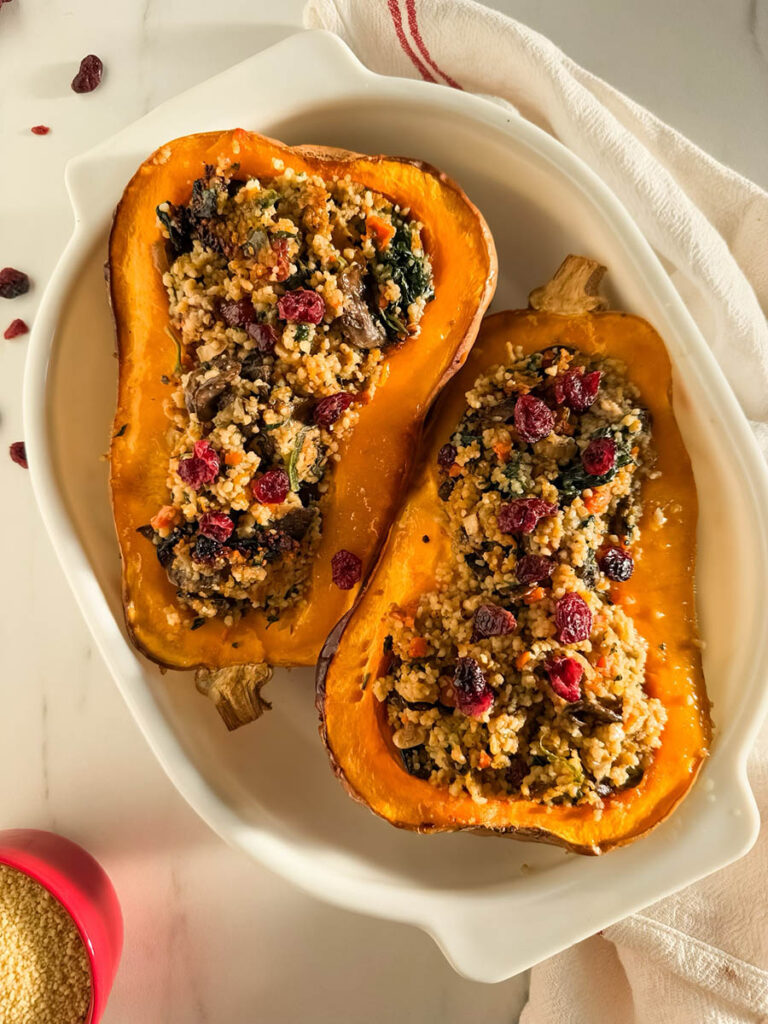 Two halves of cous cous stuffed butternut squash filled with a couscous stuffing featuring sautéed vegetables, leafy greens, and dried cranberries, presented on a white dish with a white and red-striped kitchen towel in the background, and a small red bowl of uncooked couscous nearby.