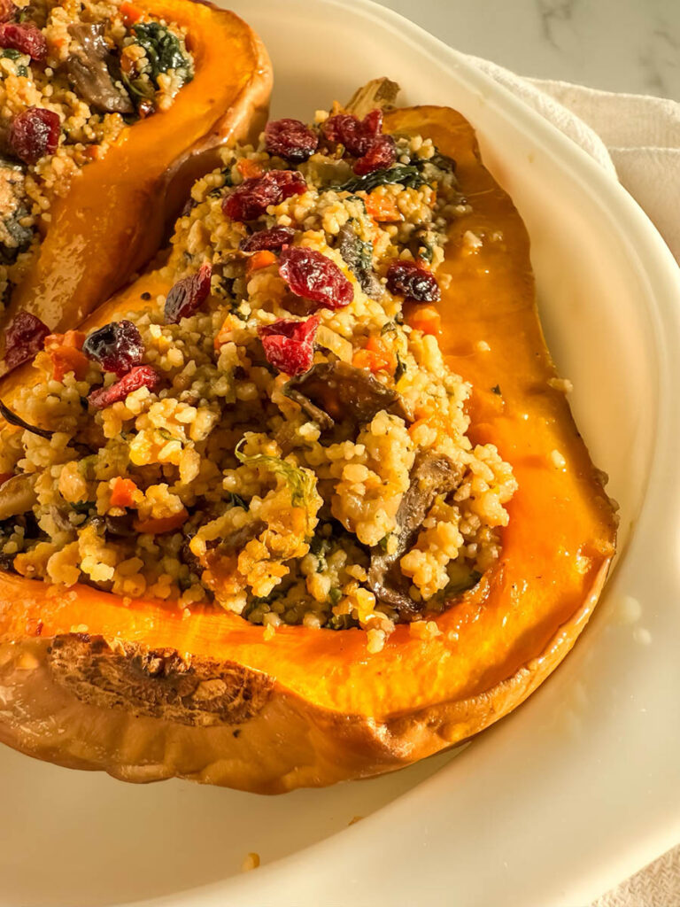 Close-up of a roasted butternut squash half filled with couscous mixed with vegetables, leafy greens, and topped with vibrant dried cranberries, served on a white plate.