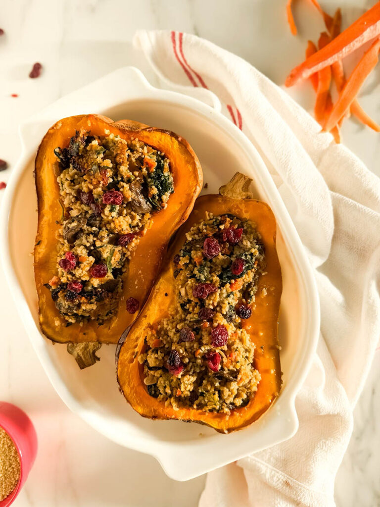 Two halves of cous cous stuffed butternut squash filled with a couscous stuffing featuring sautéed vegetables, leafy greens, and dried cranberries, presented on a white dish with a white and red-striped kitchen towel in the background, and a small red bowl of uncooked couscous nearby.