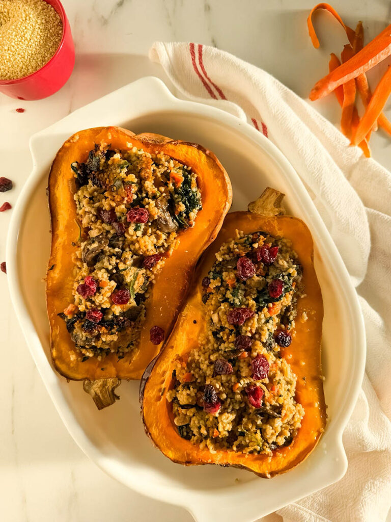 Two halves of cous cous stuffed butternut squash filled with a couscous stuffing featuring sautéed vegetables, leafy greens, and dried cranberries, presented on a white dish with a white and red-striped kitchen towel in the background, and a small red bowl of uncooked couscous nearby.