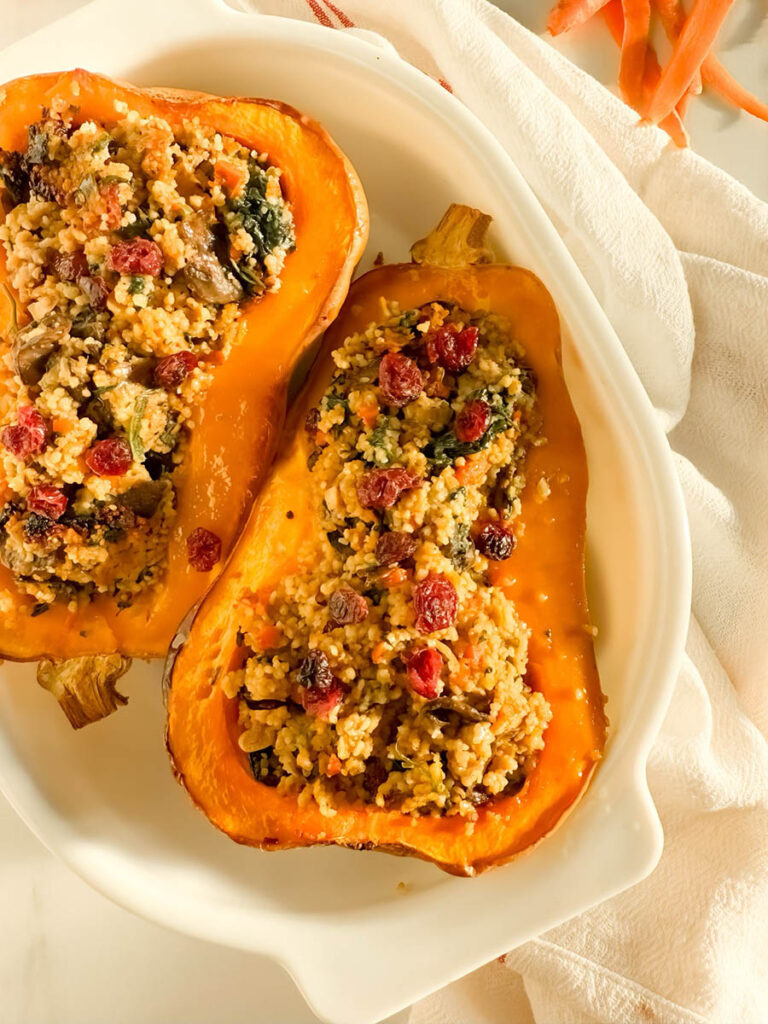 Two halves of cous cous stuffed butternut squash filled with a couscous stuffing featuring sautéed vegetables, leafy greens, and dried cranberries, presented on a white dish with a white and red-striped kitchen towel in the background, and a small red bowl of uncooked couscous nearby.