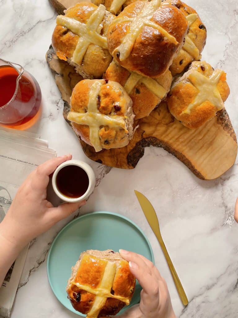 brioche Hot Cross Buns on a wooden board with a cup of coffee and a single bun on a light blue dish with a golden knife
