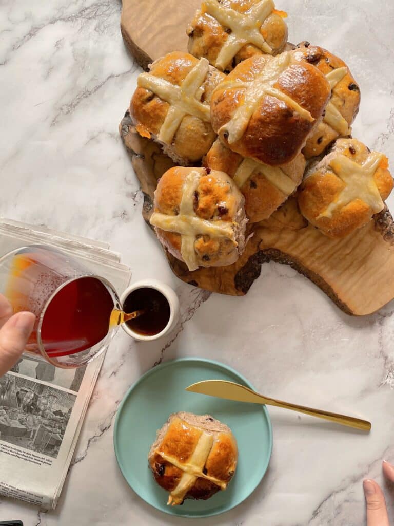 brioche Hot Cross Buns on a wooden board with a cup of coffee and a single bun on a light blue dish with a golden knife