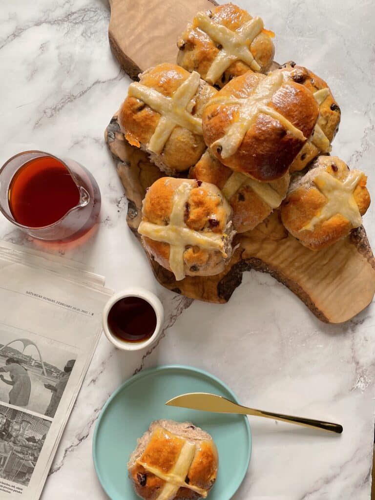 brioche Hot Cross Buns on a wooden board with a cup of coffee and a single bun on a light blue dish with a golden knife