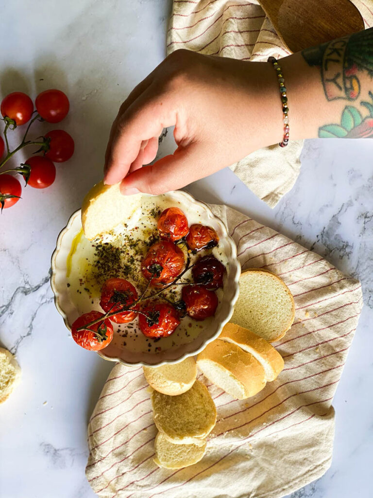 whipped feta dip in a bowl topped with roasted tomatoes and surrounded by sliced bread over a stripey napkin