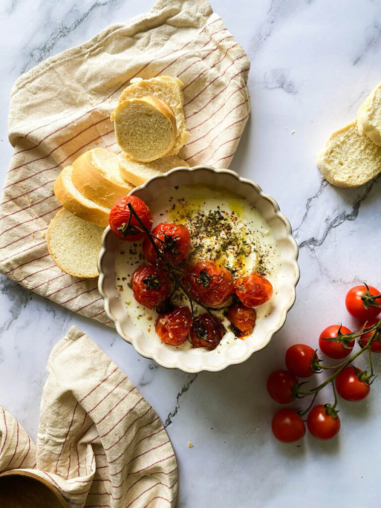 whipped feta dip in a bowl topped with roasted tomatoes and surrounded by sliced bread over a stripey napkin