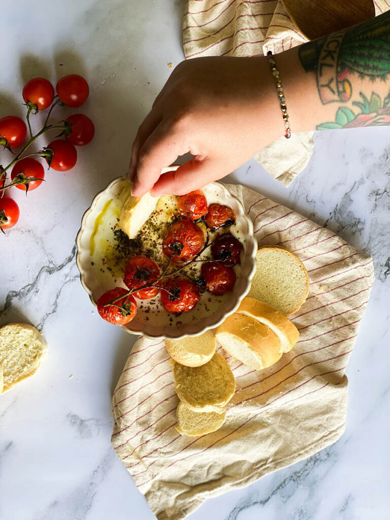 whipped feta dip in a bowl topped with roasted tomatoes and surrounded by sliced bread over a stripey napkin