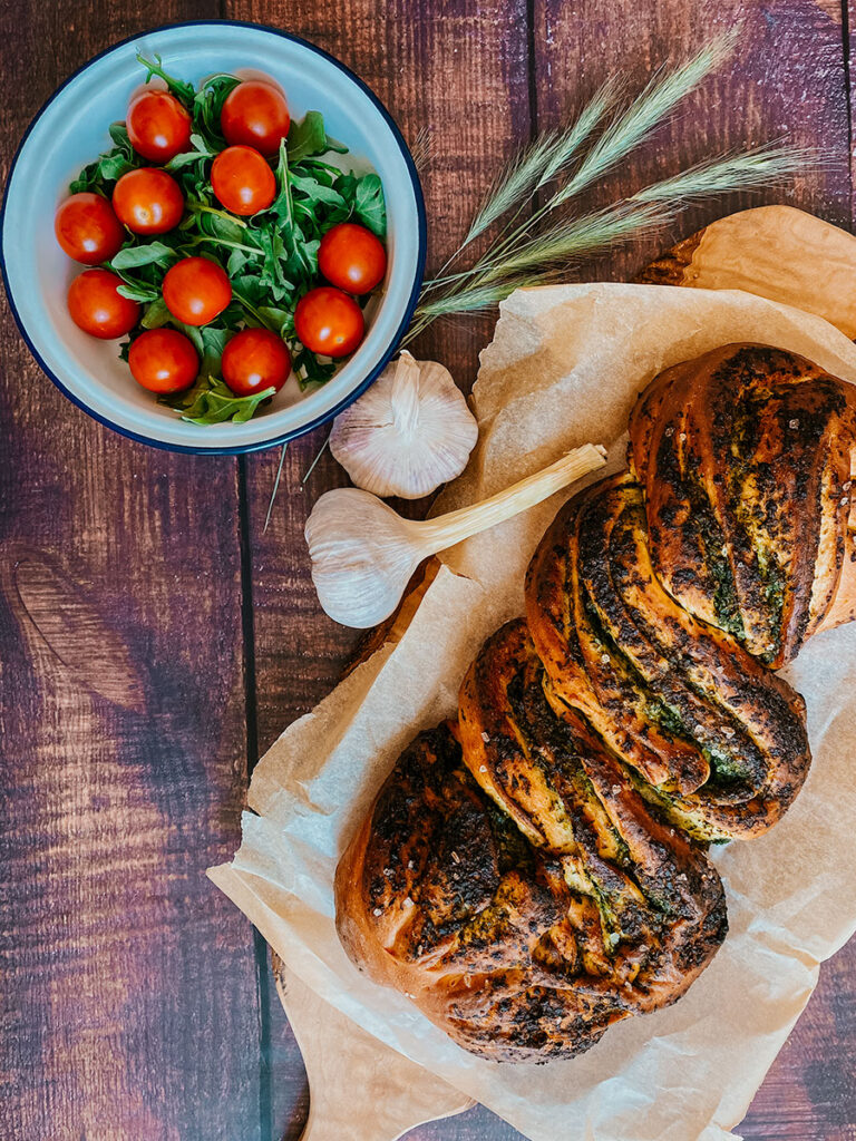Wild Garlic Bread Braided with Homemade Pesto 3 garlic bread on parchment paper, garlic and tomatoes, on a wooden background