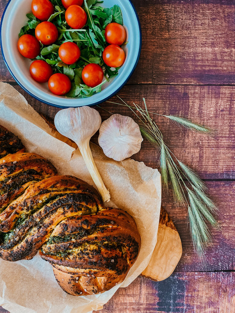 Wild Garlic Bread Braided with Homemade Pesto 1 garlic bread on parchment paper, garlic and tomatoes, on a wooden background