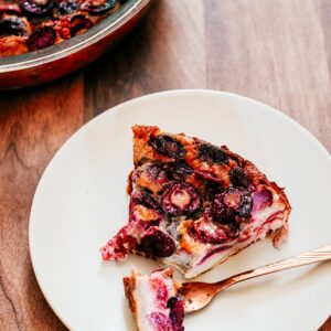 cherry clafoutis on a white plate, a piece on a fork. More clafoutis on the back