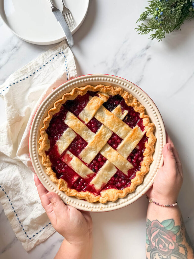 Delicious Bourbon & Mixed Berries Pie, #1 Christmas Dessert 8 Hands holding a freshly baked mixed berries pie with a golden lattice crust, placed on a marble countertop near a folded cloth, a plate, and cutlery.