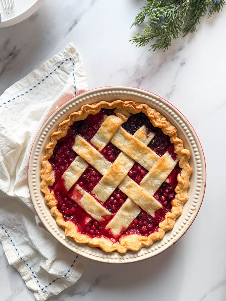 Delicious Bourbon & Mixed Berries Pie, #1 Christmas Dessert 1 A mixed berries pie with a beautifully browned lattice crust sits on a marble surface with a folded white cloth nearby, viewed from above.