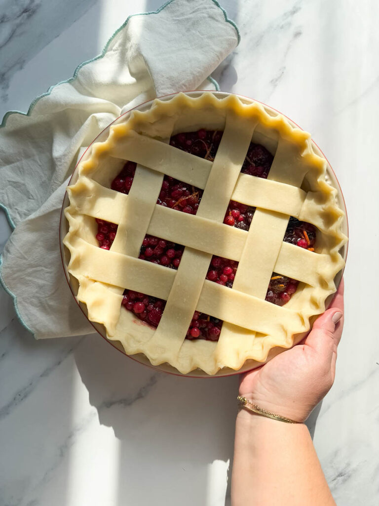 Delicious Bourbon & Mixed Berries Pie, #1 Christmas Dessert 4 A hand holding an unbaked mixed berries pie with a lattice crust, ready for the oven, on a bright surface next to a folded cloth.