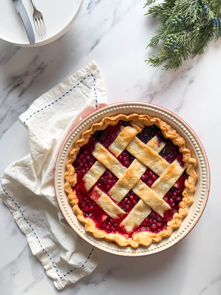Delicious Bourbon & Mixed Berries Pie, #1 Christmas Dessert 6 A mixed berries pie with a beautifully browned lattice crust sits on a marble surface with a folded white cloth nearby, viewed from above.