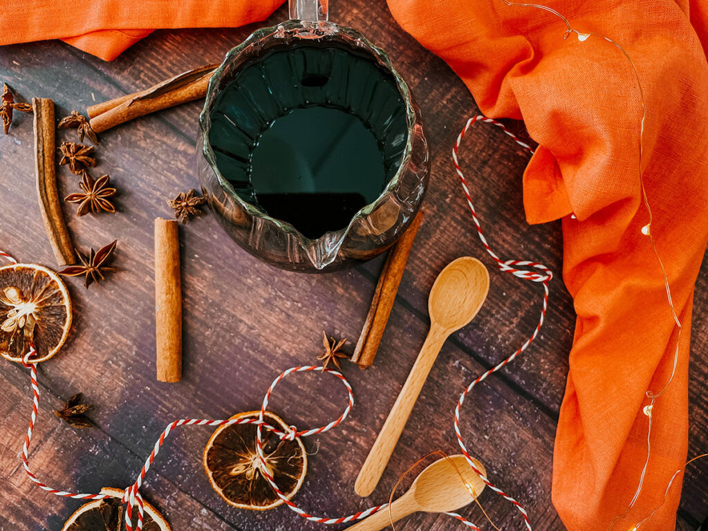 Gingerbread Syrup Recipe For A Delicious Christmassy Gingerbread Latte 3 Overhead view of a glass pitcher filled with gingerbread syrup surrounded by dried orange slices, cinnamon sticks, star anise, and wooden spoons on a rustic wooden table. Bright orange fabric and twine add a festive, warm touch to the scene.