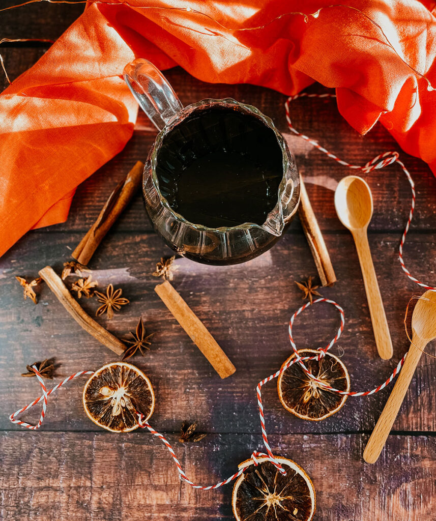 Gingerbread Syrup Recipe For A Delicious Christmassy Gingerbread Latte 1 Overhead view of a glass pitcher filled with gingerbread syrup surrounded by dried orange slices, cinnamon sticks, star anise, and wooden spoons on a rustic wooden table. Bright orange fabric and twine add a festive, warm touch to the scene.