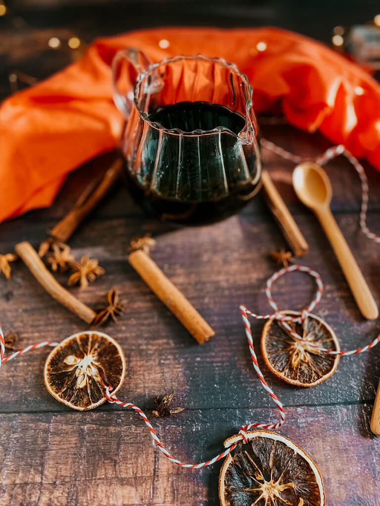 Gingerbread Syrup Recipe For A Delicious Christmassy Gingerbread Latte 4 Overhead view of a glass pitcher filled with gingerbread syrup surrounded by dried orange slices, cinnamon sticks, star anise, and wooden spoons on a rustic wooden table. Bright orange fabric and twine add a festive, warm touch to the scene.