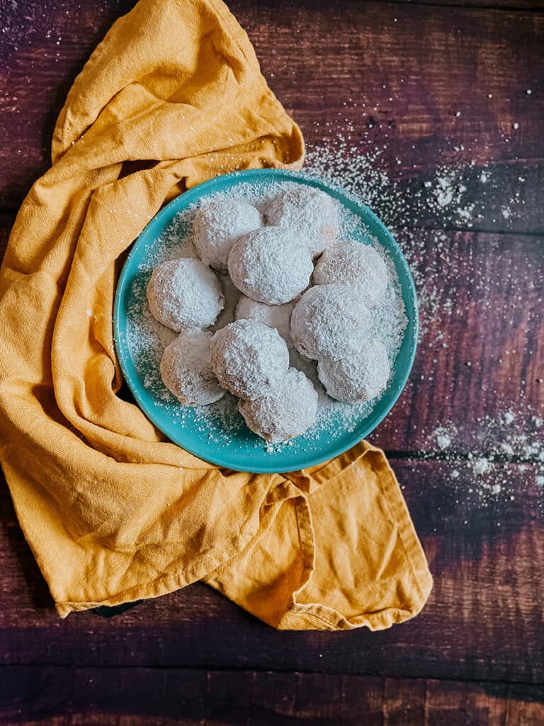 Kourabiethes, Delicious & Vegan Greek Cookies 1 A plate of Greek kourabiethes cookies dusted with powdered sugar sits on a teal plate, placed on a rustic wooden surface. The cookies are arranged next to a mustard-yellow cloth, with a light dusting of sugar scattered around, creating a cozy, homemade feel.