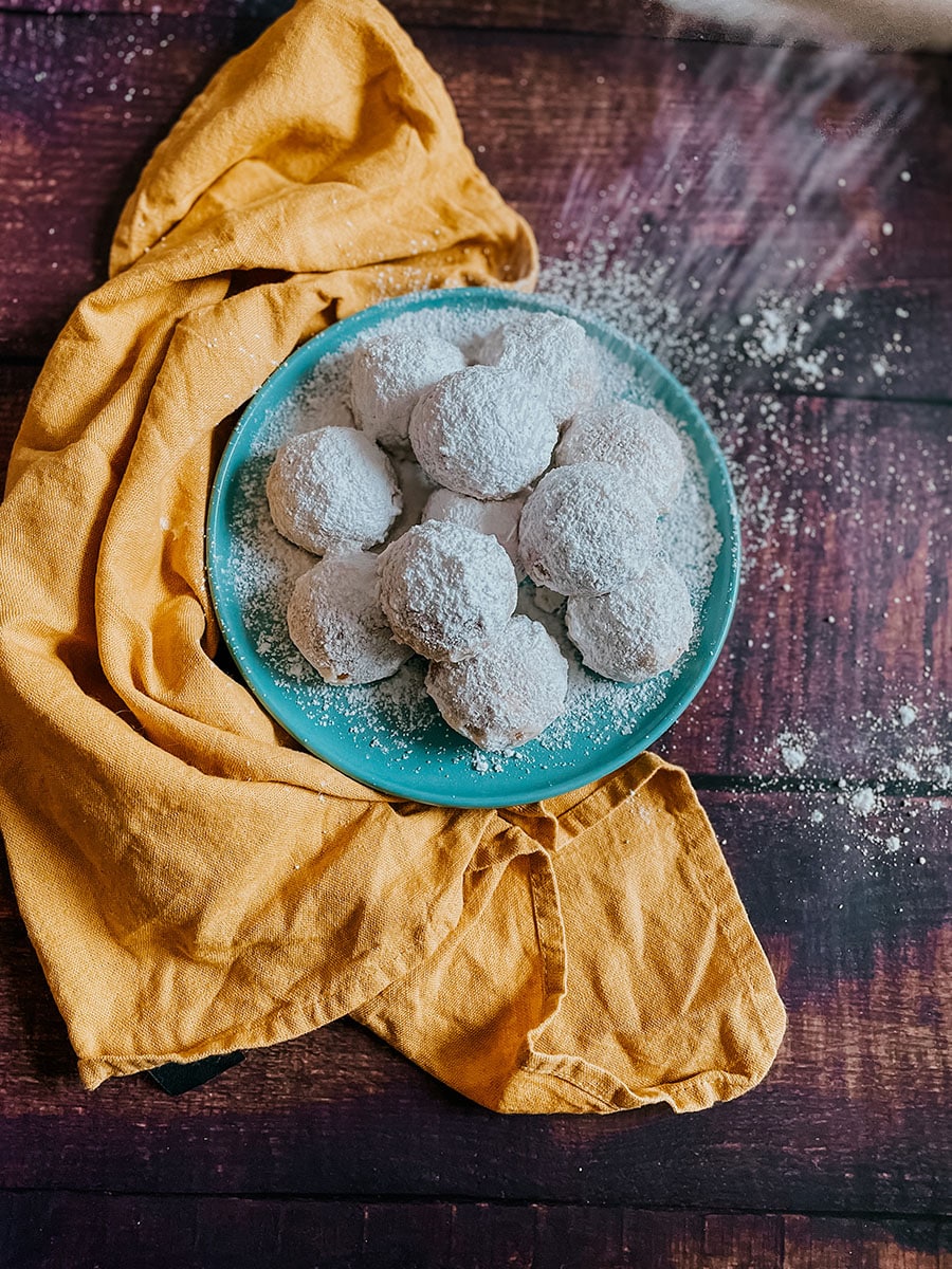 Kourabiethes, Delicious & Vegan Greek Cookies 3 A plate of Greek kourabiethes cookies dusted with powdered sugar sits on a teal plate, placed on a rustic wooden surface. The cookies are arranged next to a mustard-yellow cloth, with a light dusting of sugar scattered around, creating a cozy, homemade feel.