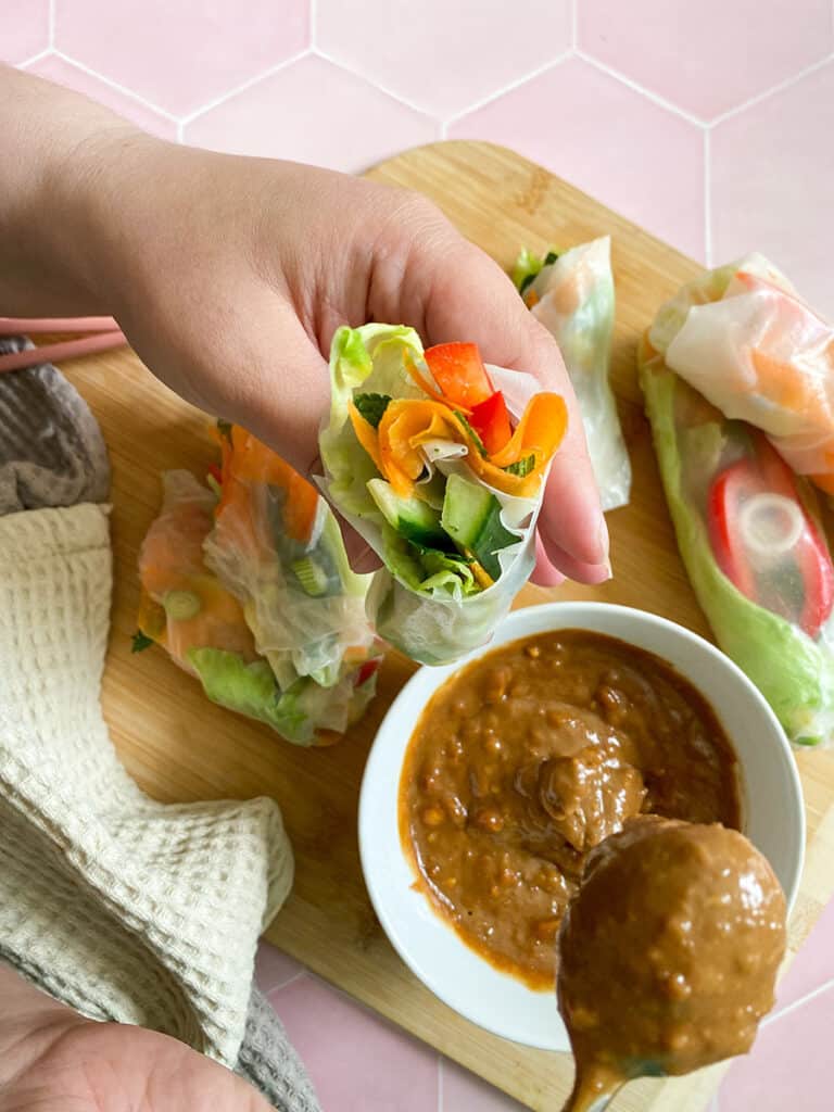 Vegan Rice Paper Rolls With Yummy Peanut Dipping Sauce 5 vegan rice paper rolls with a peanut dipping sauce in a white bowl with a spoon on a wooden board