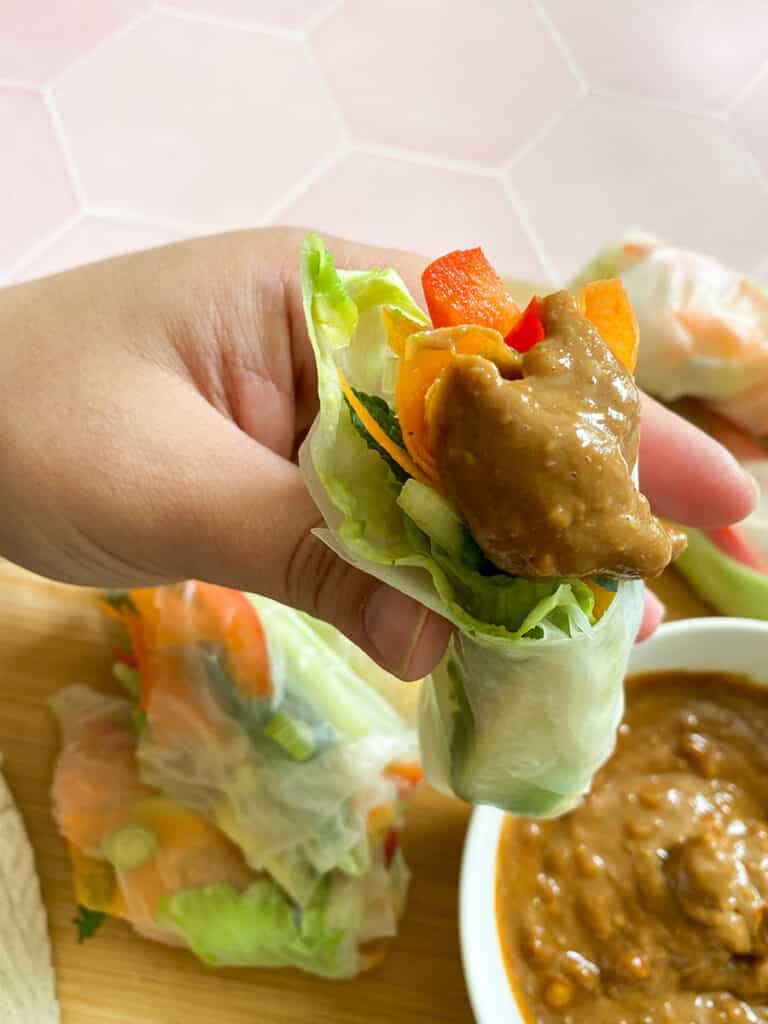 Vegan Rice Paper Rolls With Yummy Peanut Dipping Sauce 6 vegan rice paper rolls with a peanut dipping sauce in a white bowl with a spoon on a wooden board
