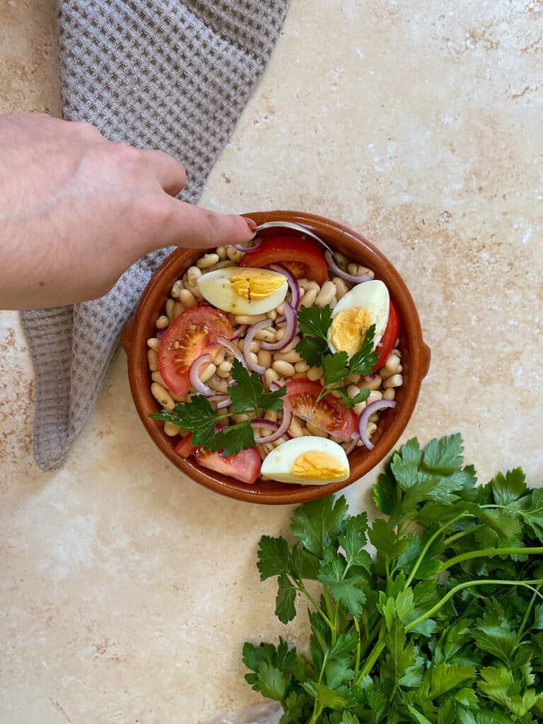 10 Minutes Summer Butter Beans Salad 4 hand with a spoon holding a tomato, under a bowl of butter beans salad, a towel and parsley