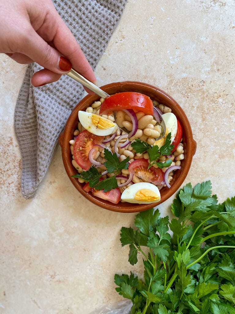 10 Minutes Summer Butter Beans Salad 5 hand with a spoon holding a tomato, under a bowl of butter beans salad, a towel and parsley
