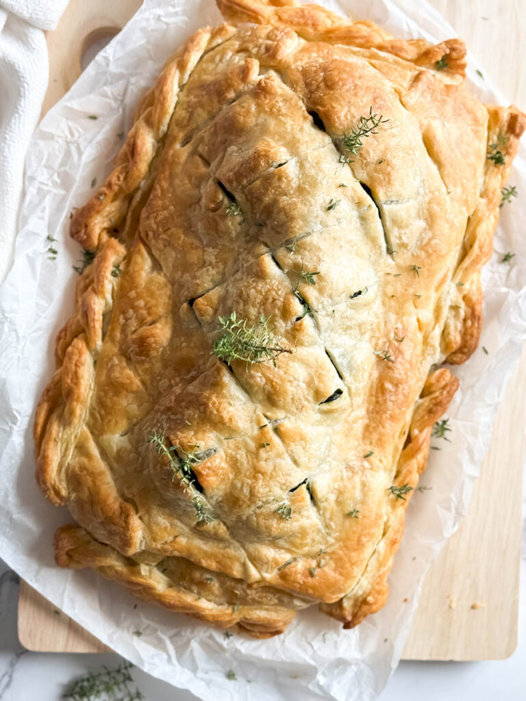 A golden-brown vegan mushroom Wellington on a sheet of parchment paper, garnished with fresh thyme, with a rustic kitchen towel and onion peels in the background.