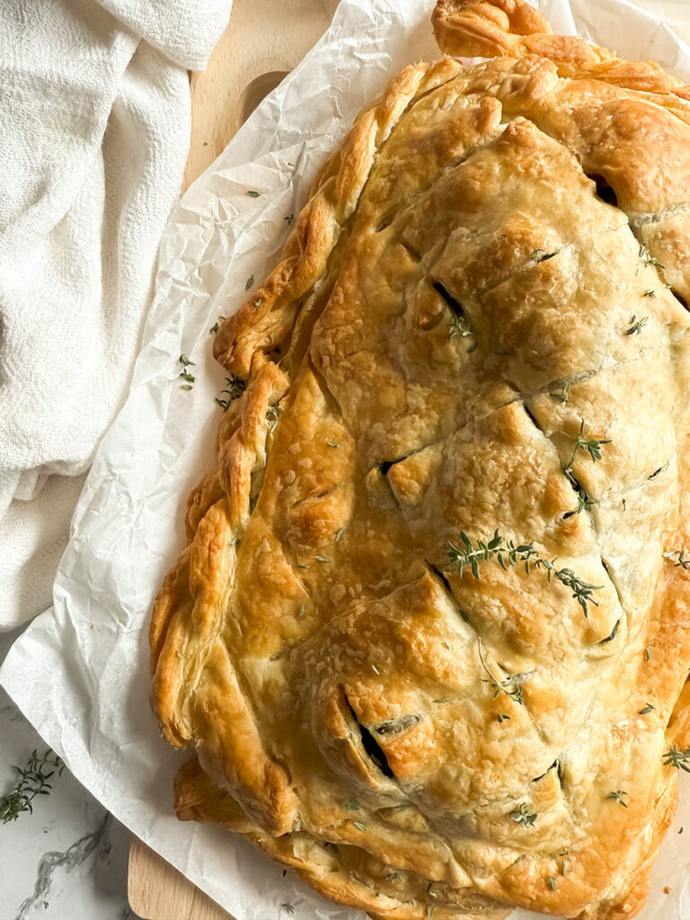 A golden-brown vegan mushroom Wellington on a sheet of parchment paper, garnished with fresh thyme, with a rustic kitchen towel and onion peels in the background.