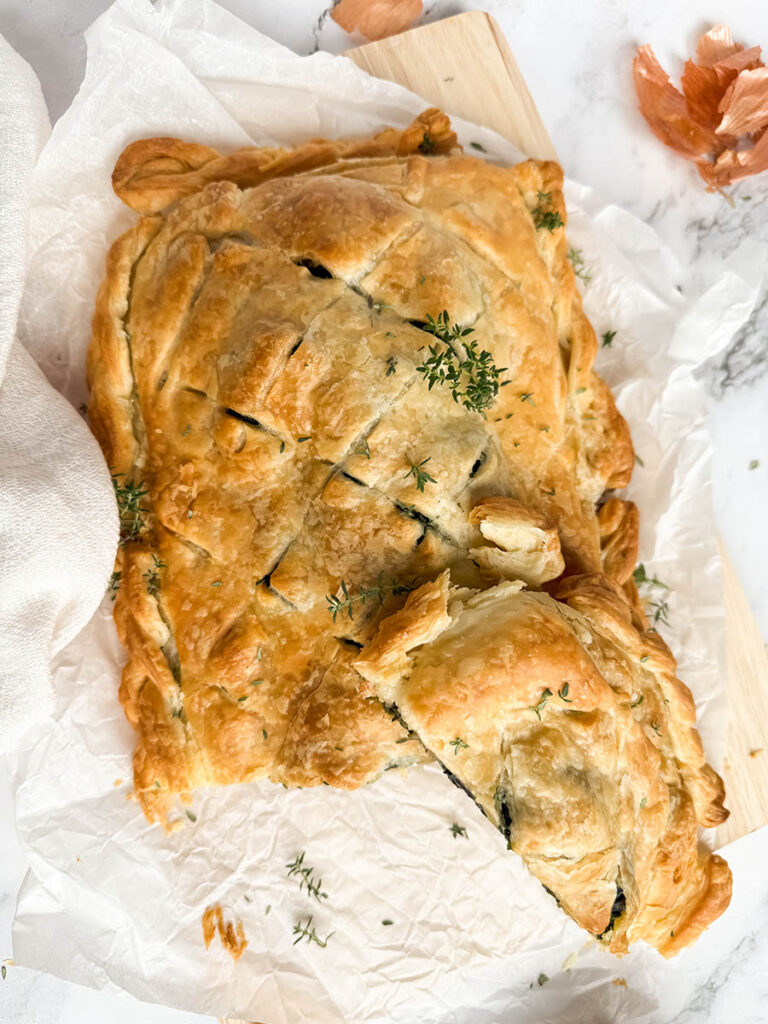 A sliced vegan mushroom Wellington, showing the flaky crust and the interior, surrounded by bits of thyme and onion peels on a wooden cutting board.