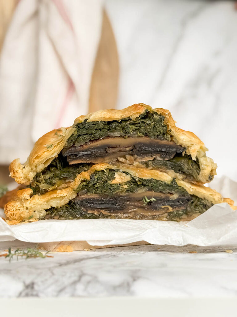 A close-up of a cross-section of the vegan mushroom Wellington, revealing layers of mushrooms and spinach encased in a flaky pastry crust, with thyme sprinkled on top.