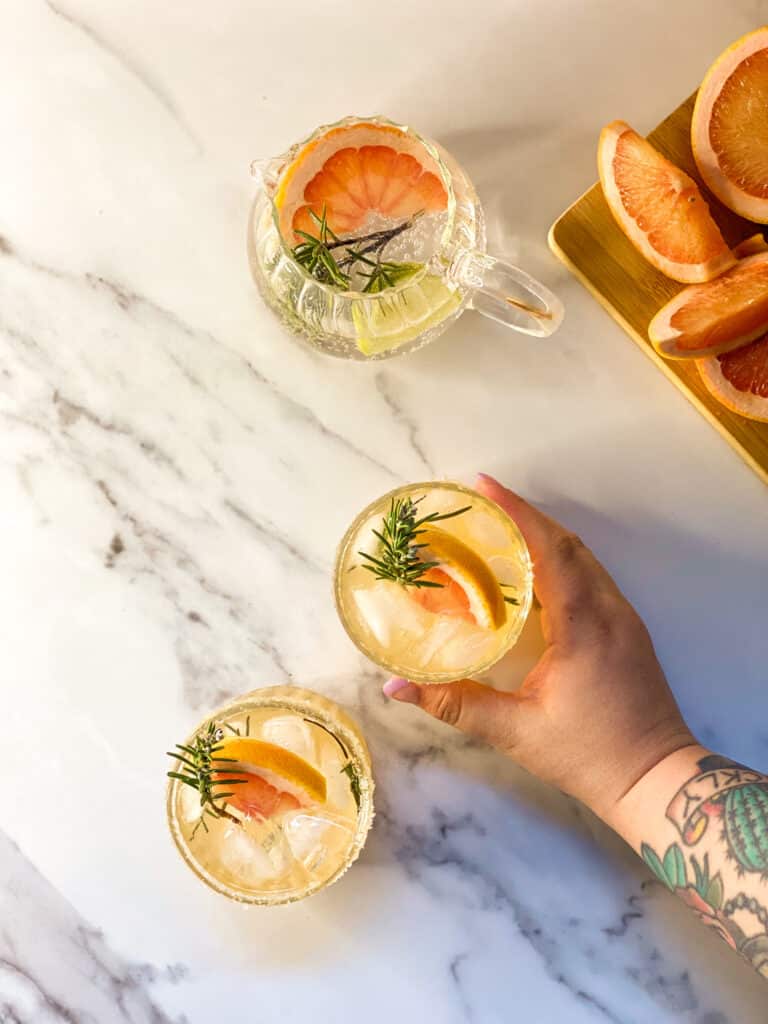 Amazing Sparkling Grapefruit Mocktail Recipe 2 two glasses containing a grapefruit mocktail with rosemary, a hand reaching for one