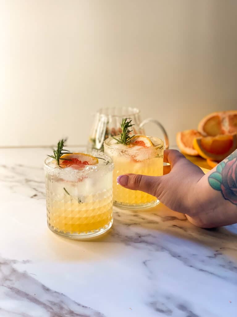 Amazing Sparkling Grapefruit Mocktail Recipe 6 two glasses containing a grapefruit mocktail with rosemary, a hand holding a glass