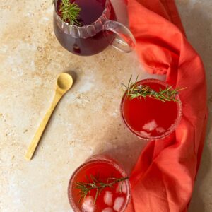 5 Minutes Love Potion Cocktail Recipe 5 two glasses with the love potion drink with a rosemary twig as garnish, a glass jug of juice on the back, a wooden spoon on the left and a coral red napkin on the right