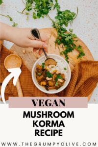 Vegan Mushroom Korma, The #1 Best Recipe 7 A hand holding a spoon scoops mushroom korma from a bowl of rice, surrounded by fresh cilantro sprigs. The bowl sits on a wooden cutting board with a matching wooden spoon and a rust-colored towel, creating a cozy presentation.