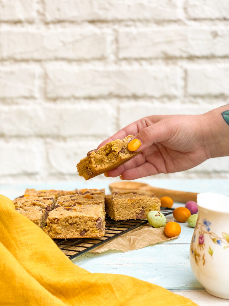 mini eggs blondies on a cookie rack, mini eggs on the side, a hand reaching for one