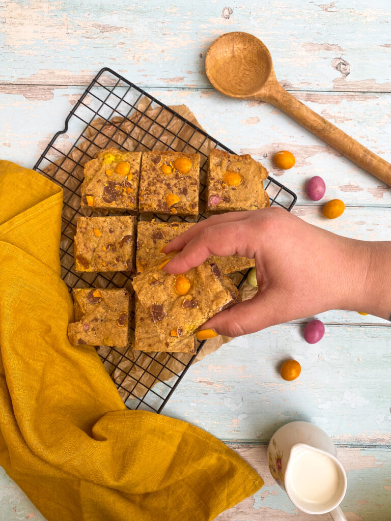 mini eggs blondies on a cookie rack, mini eggs on the side, a hand reaching in 