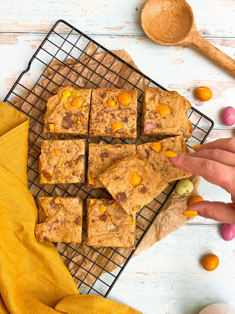  mini eggs blondies on a cookie rack, a hand reaching for one, a wooden spoon on the side