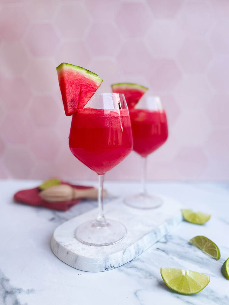 watermelon tequila spritzer served in two wine glasses, sitting on a marble slate with a slice of watermelon each and some lime wedges scattered around