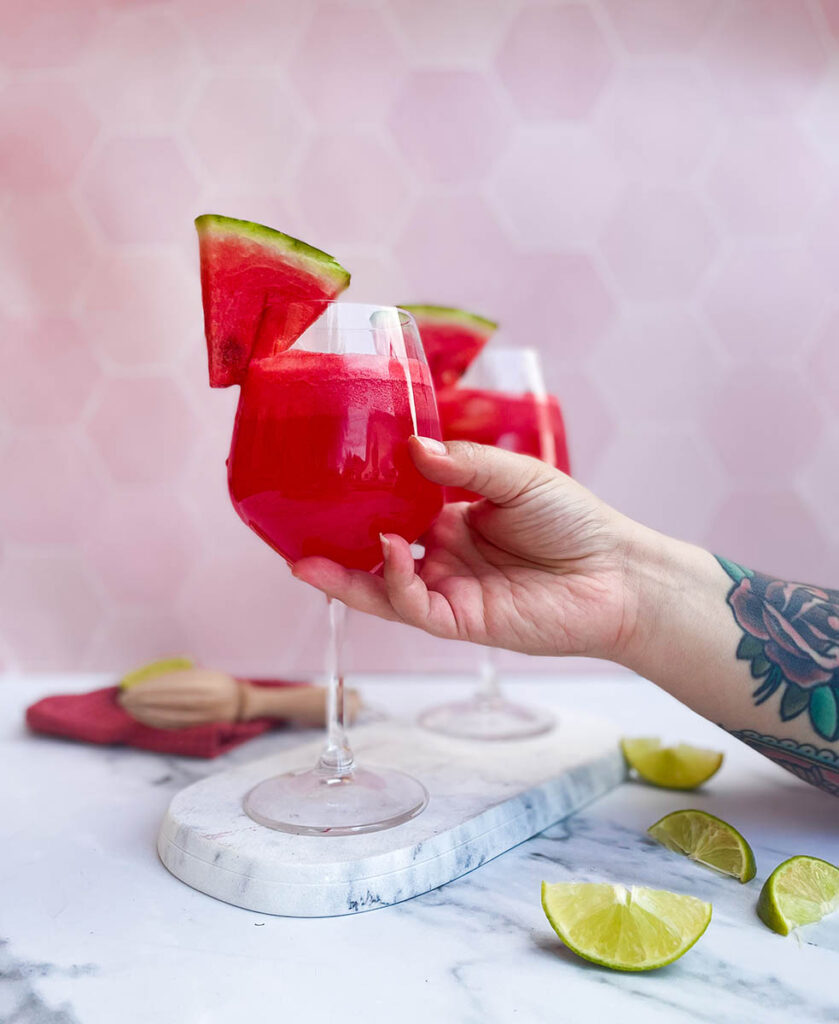 watermelon tequila spritzer served in two wine glasses, one of them being held by a hand, sitting on a marble slate with a slice of watermelon each and some lime wedges scattered around