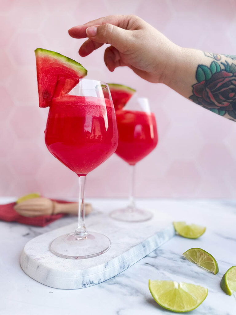 watermelon tequila spritzer served in two wine glasses, sitting on a marble slate with a slice of watermelon each and some lime wedges scattered around