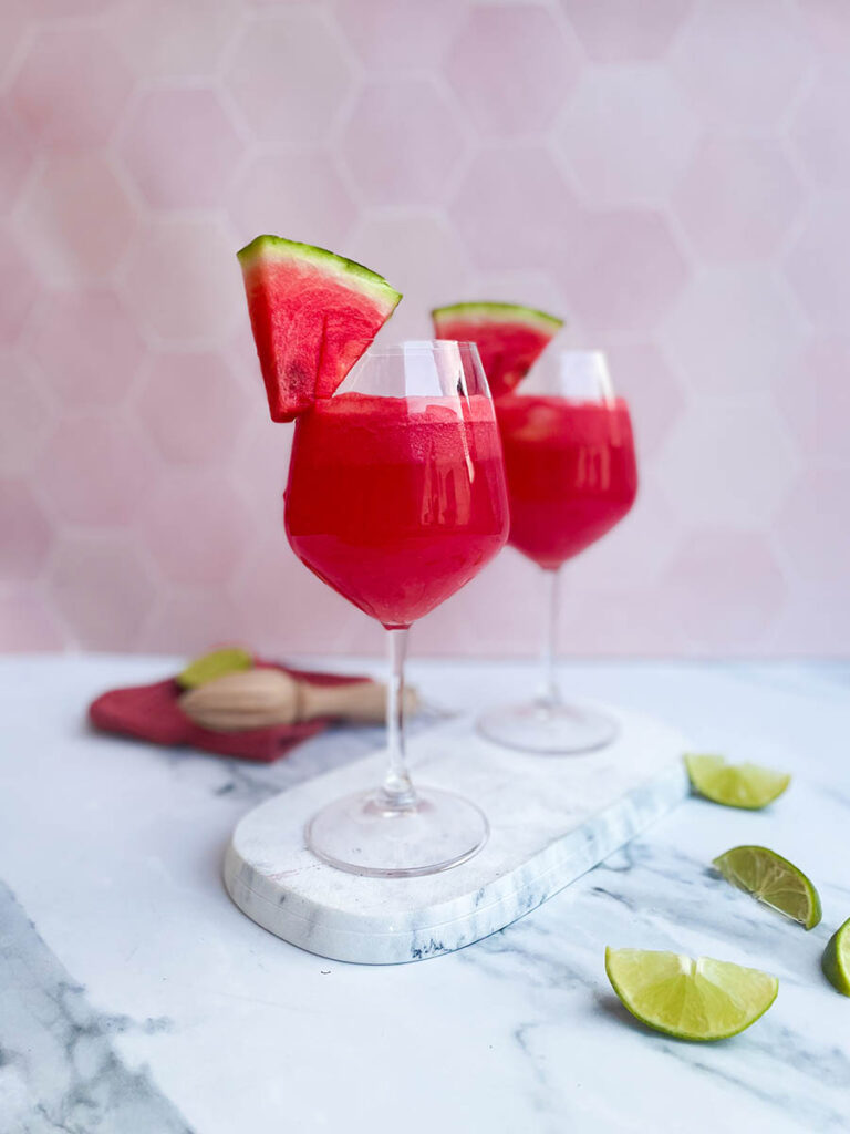 watermelon tequila spritzer served in two wine glasses, sitting on a marble slate with a slice of watermelon each and some lime wedges scattered around
