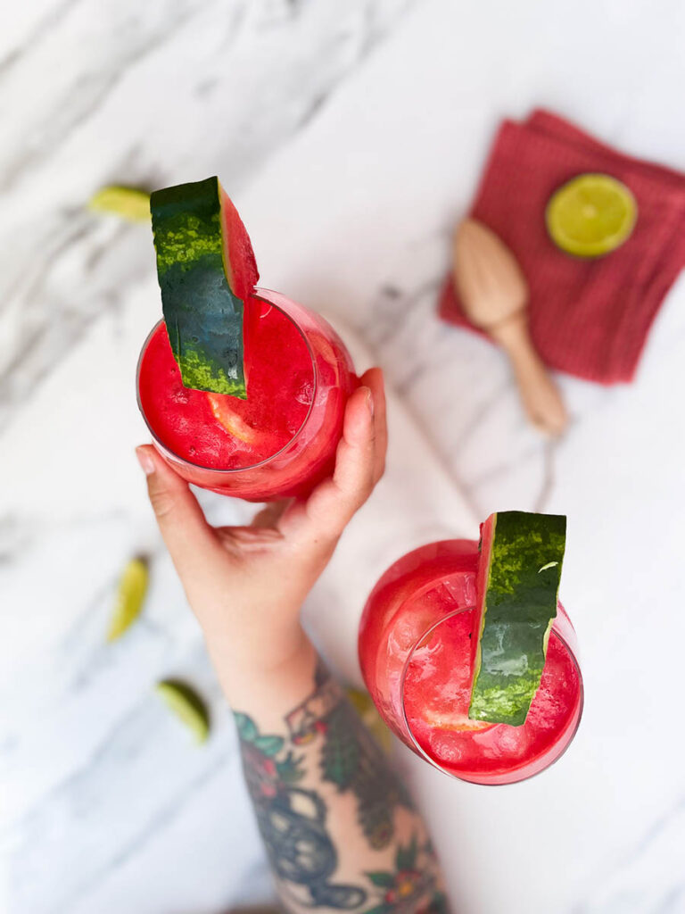 watermelon tequila spritzer from above served in two wine glasses, sitting on a marble slate with a slice of watermelon each and some lime wedges scattered around