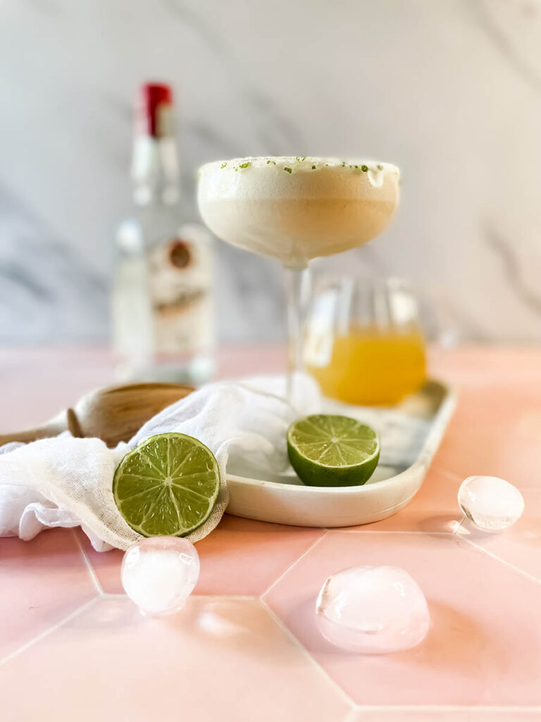 pineapple coconut margarita glass on a marble tray, with ice and lime on the foreground, a white napkin, a wooden juicer and a jug of juice and tequila bottle on the background